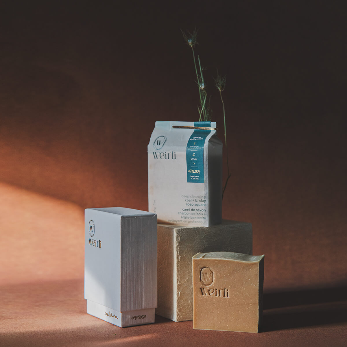 Deep cleansing coal and bentonite soap square in its box, in it's interior packaging, and unpackaged soap. On a warm background with twigs peaking through a concrete cube. 