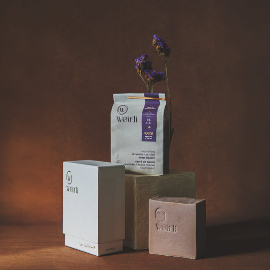 Weirli products arranged on a wooden surface against a warm brown backdrop: a white rectangular box, a kraft paper bag with a purple label, and a rectangular bar of soap in a muted pinkish-beige container. A small vase with dried purple flowers sits behind the bag. The products are raw soap, soap in an interior packaging, and the outer box.